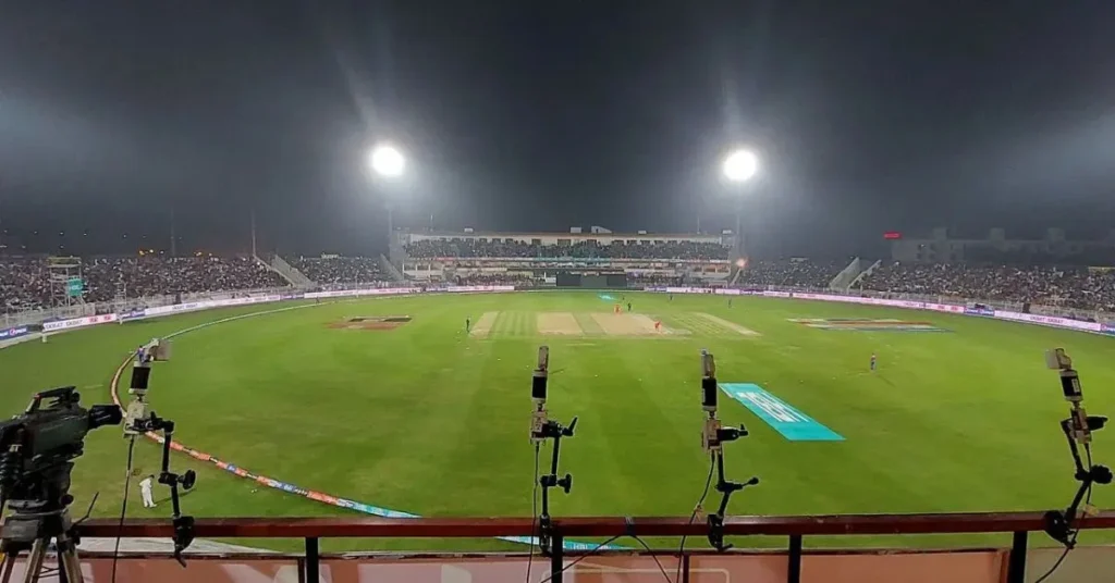 The image shows Rawalpindi Cricket Stadium illuminated at night under bright floodlights. The stadium is filled with a large crowd of spectators, and a cricket match is in progress on the well-maintained green field. The image captures a vibrant atmosphere with cameras positioned in the foreground for broadcasting.