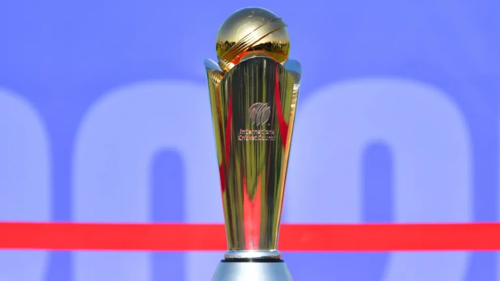 The image shows the ICC Champions Trophy, which has a sleek golden and silver design featuring a globe-like top. The ICC logo and "International Cricket Council" text are engraved on the front. The trophy is displayed against a blue background with a red stripe across it.