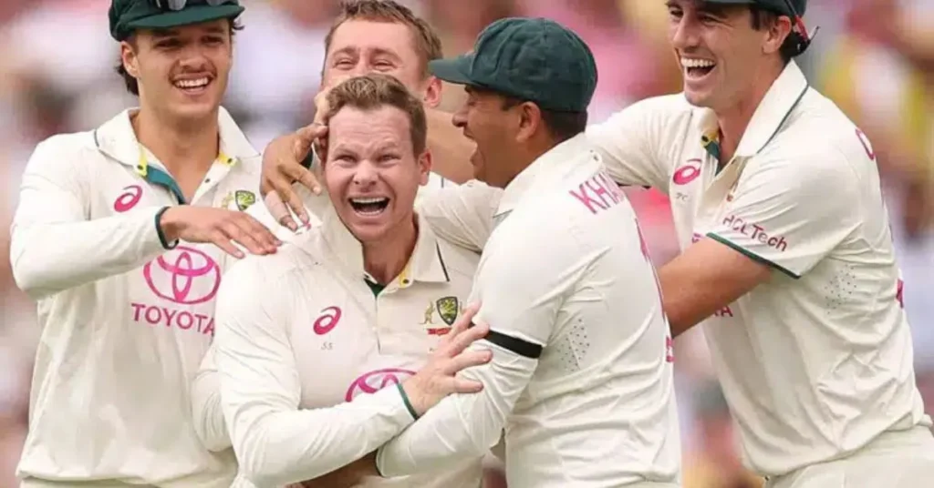 Australian cricket players celebrating together on the field during a Test match, with one player, visibly thrilled, at the center of the group hug while others cheer and smile.